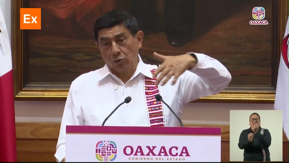 A man in a traditional white guayabera shirt speaks at an OAXACA (Gobierno del Estado) podium with microphones, while a sign language interpreter appears in a picture-in-picture window in the bottom right corner; an orange 'Ex' logo is in the top left.