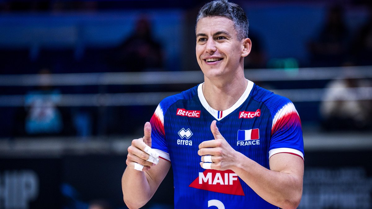 Jenia Grebennikov, one of volleyball's most decorated liberos, smiles and gives a double thumbs-up while wearing the blue France national team jersey (featuring "errea," "MAIF," and "Betclic" sponsorships) before joining Japan's Wolfdogs Nagoya for the SV League season.