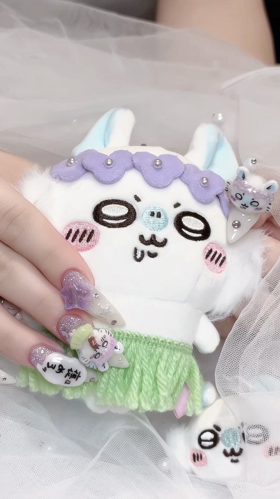 A close-up of a person's hands with pastel, decorated nails holding a cute plush toy with purple and blue accents, worn with a sheer white tulle garment.