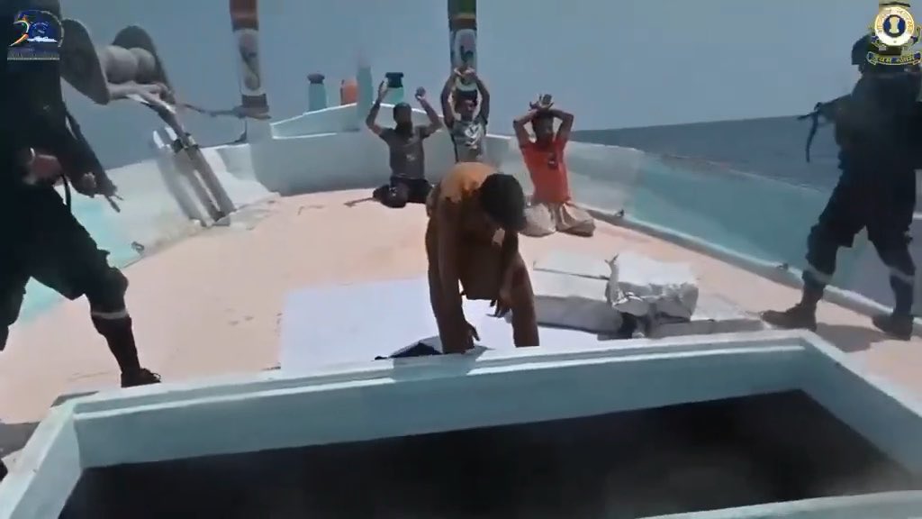 Several men kneel with hands behind their heads on the deck of a boat at sea while armed personnel stand guard over them.