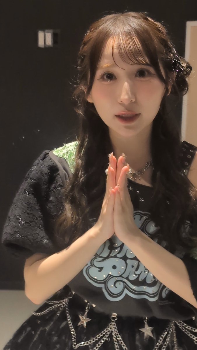 A young woman with long dark hair presses her hands together in a prayer-like gesture, wearing a dark outfit with a 'CONRN' graphic and a star-patterned skirt.