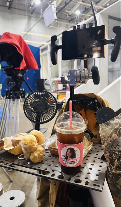 A behind-the-scenes film production setup featuring camera gear, a small fan, iced coffee in a branded cup with a woman's photo, and pastries on a perforated metal table.