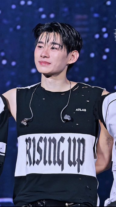 A young man with dark hair wearing a black and white shirt with "RISING UP" printed in gothic-style text, alongside the word "ATLAS".