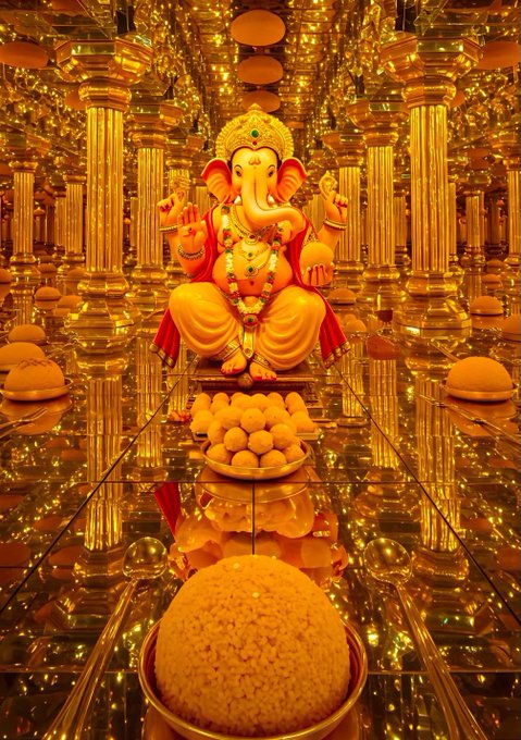An ornate golden statue of the elephant-headed deity Ganesha sits in a mirror-lined temple hall adorned with columns and lit candles, with plates of round yellow sweets (ladoos) placed before it.