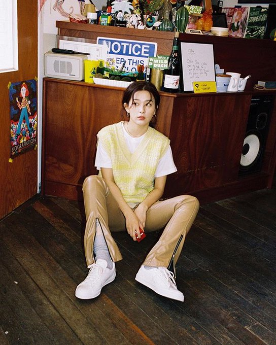 A young woman sitting on a wooden floor in a yellow knit vest, white tee, and beige zip pants with white sneakers, in a room with wooden cabinets and various decorative items.