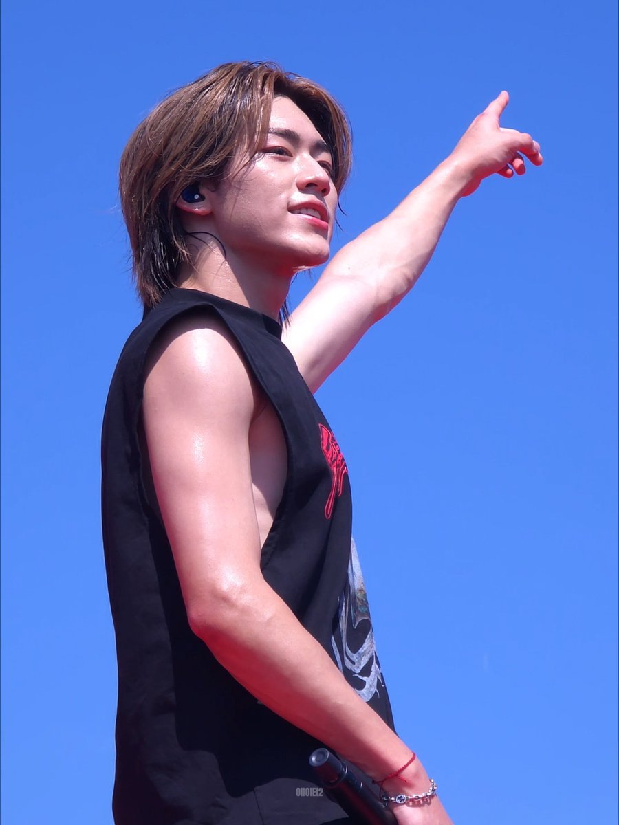 A performer with shoulder-length brown hair, wearing a black sleeveless top and in-ear monitors, points dramatically upward against a clear blue sky during an outdoor performance.