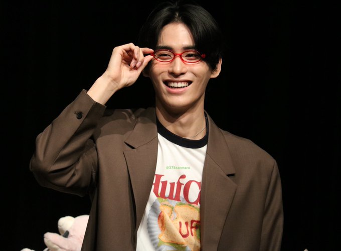 A smiling young man with glasses playfully holds the frame of his red glasses, wearing a brown blazer over a t-shirt featuring "tulca" text and "378iamneru".