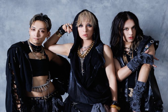 A promotional photo of three members of the Japanese band Xaa Xaa posed against a gray backdrop, wearing edgy black outfits with gold chains and gothic styling.