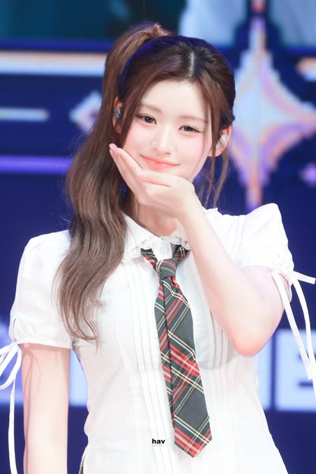 A young woman with long brown hair in a side ponytail poses with a hand near her chin, wearing a white blouse with a plaid necktie and a headband reading 'nav' on the shirt.