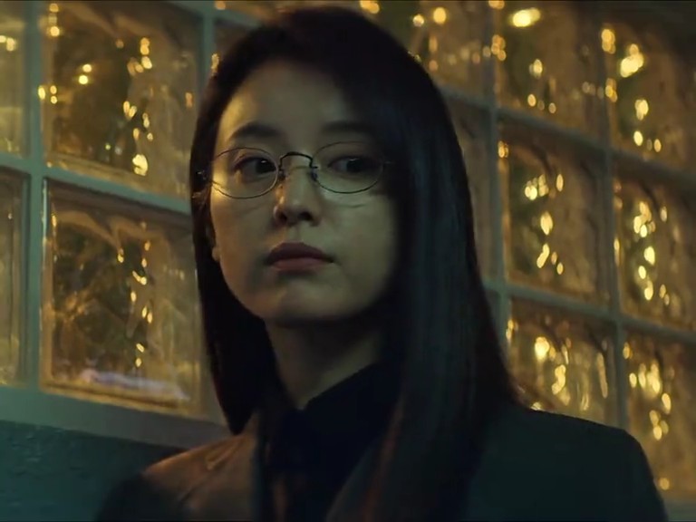 A close-up of a woman with long dark hair and round wire-rimmed glasses, wearing a dark top. She has a serious expression and is positioned in front of a textured glass block wall illuminated by warm, golden light.