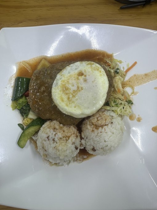 A plated meal featuring a hamburger patty topped with a fried egg, served alongside rice balls, stir-fried noodles, and vegetables with sauce on a white plate.
