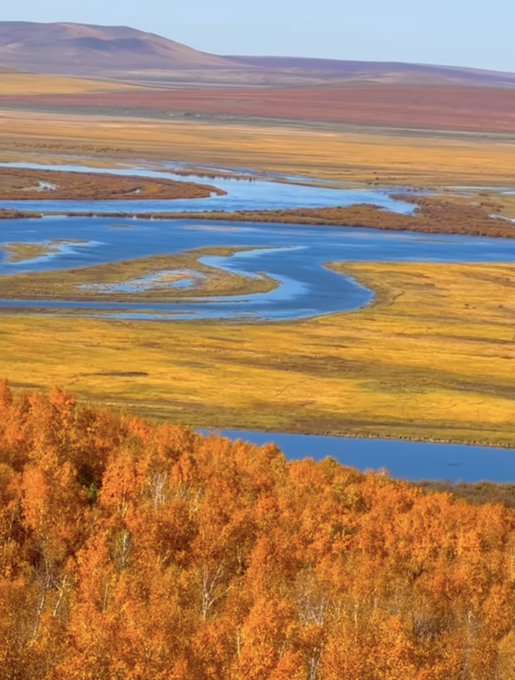 A scenic landscape features a winding blue river cutting through golden grasslands, autumn foliage in the foreground, and rolling hills in the distance under a clear sky.
