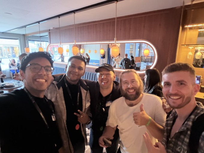 Group selfie of five smiling men posing together at a casual restaurant or event venue.