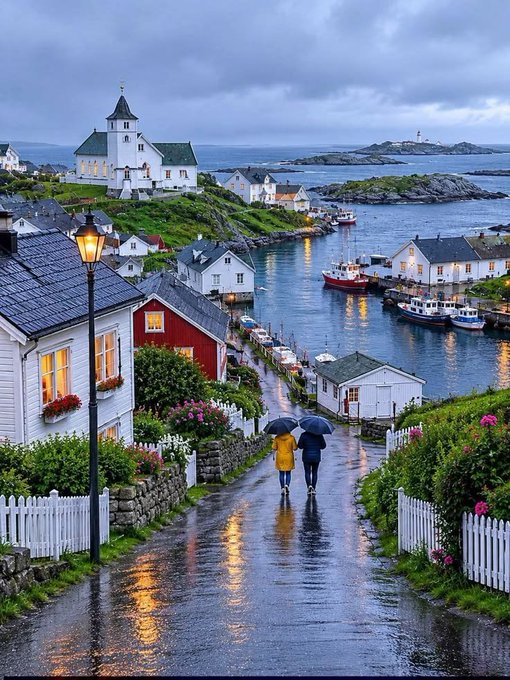 A scenic coastal village with a white church, colorful houses, boats in the harbor, and two people walking with umbrellas down a wet cobblestone path on a rainy evening.