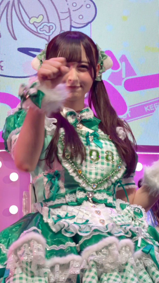 A young performer with long dark pigtails wears an elaborate green and white gingham idol dress with ruffles and bows, posing playfully toward the camera on a colorful stage backdrop. The image reflects the post's sentiment of missing "Noa-chan," likely referring to the idol member in the photo, whose cheerful expression and cute outfit evoke fond nostalgia for fans.