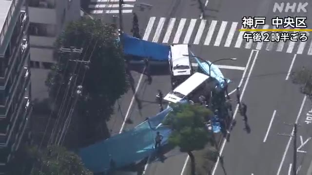 An NHK aerial news clip from Kobe City (神戸中央区), captured around 2:30 PM (午後2時半ごろ), shows what appears to be a crime scene with blue tarps covering the sidewalk near a crosswalk, consistent with coverage of a possible serial shooting incident where two adult men were injured and an elderly suspect was detained.