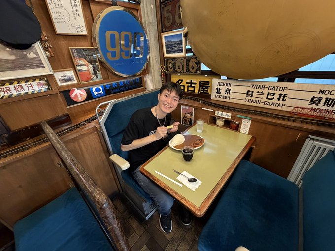 A young man sits in a vintage-style restaurant booth with eclectic decor, eating a meal with chopsticks and a glass of drink on a green table.