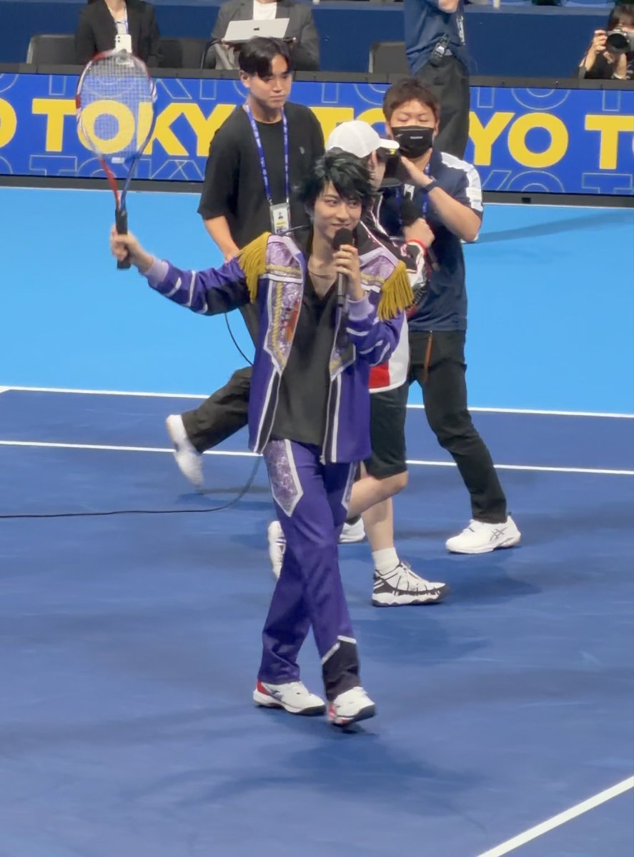 A man in an elaborate purple and gold costume holds a tennis racket on a blue court with "TOKYO" signage in the background, while staff members stand nearby. The theatrical performance setup aligns with the post about Imai Ryōma's genuine delivery and the crowd's enthusiastic response during what appears to be a tennis-themed event.