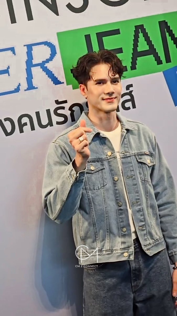 A young man with wavy brown hair, wearing a light blue denim jacket, makes a finger heart gesture at a fan event with a "SINGHA" and Thai text banner behind him; watermarked "CM 2 CONNECT".