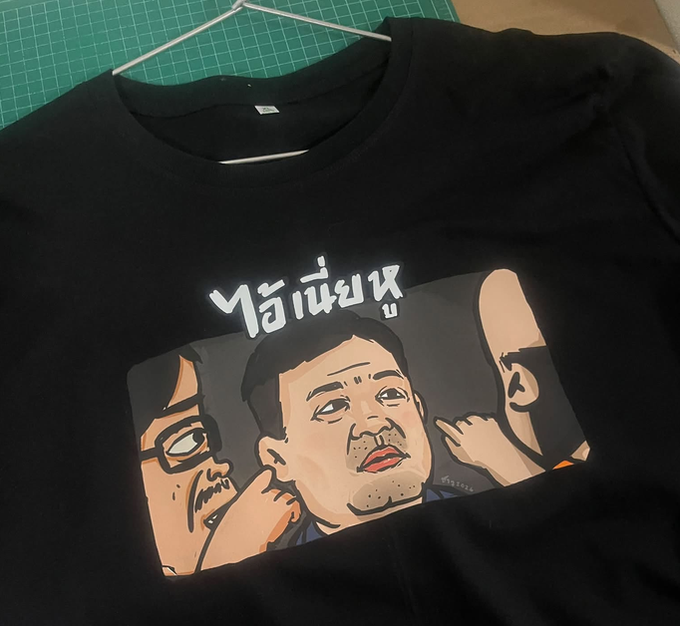 A black T-shirt hanging on a wire hanger on a green cutting mat, featuring an anime-style cartoon illustration of three men and Thai script reading "ไอ้เน่คุง".