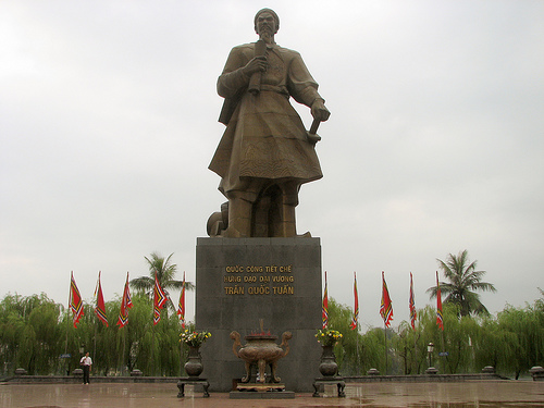 A large bronze statue of a historical figure in traditional Vietnamese attire standing on a stone pedestal with an incense burner, surrounded by red flags, with the inscription 'ĐƯỚC CÔNG THẤT CHẾ UẤT ĐẮP ĐẠI VƯƠNG TRAN QUỐC TUẤN'.