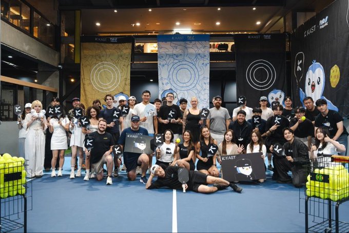 A large group photo of people posing on a blue tennis court at a KRAST event, holding K and KRAST signs with tennis balls in baskets.