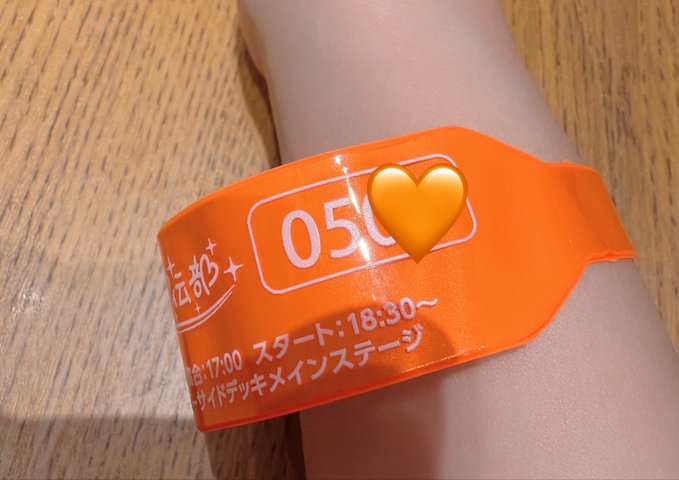A bright orange event wristband on a person's wrist, displaying the number "05" and Japanese text including "スタート:18:30" (start time) with a yellow heart emoji obscuring part of the wristband.