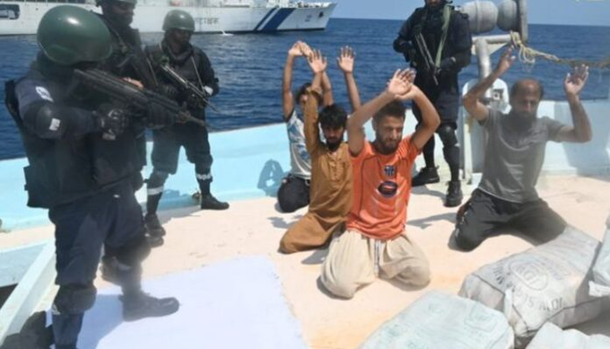 Armed personnel in tactical gear standing guard over kneeling men with their hands raised on what appears to be a ship deck, with packages visible in the foreground.