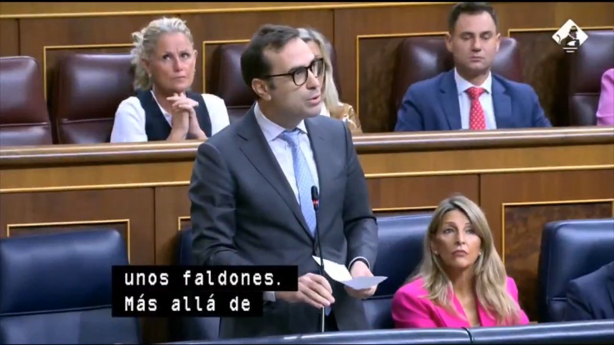 A man in a dark suit and glasses speaks at a podium in what appears to be a parliamentary chamber, with Spanish subtitles reading 'unos faldones. Más allá de' visible on screen.