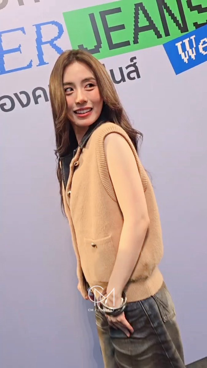 A young woman wearing a tan knit vest over a black top smiles while posing in front of a 'JEANS' event backdrop with Thai text.