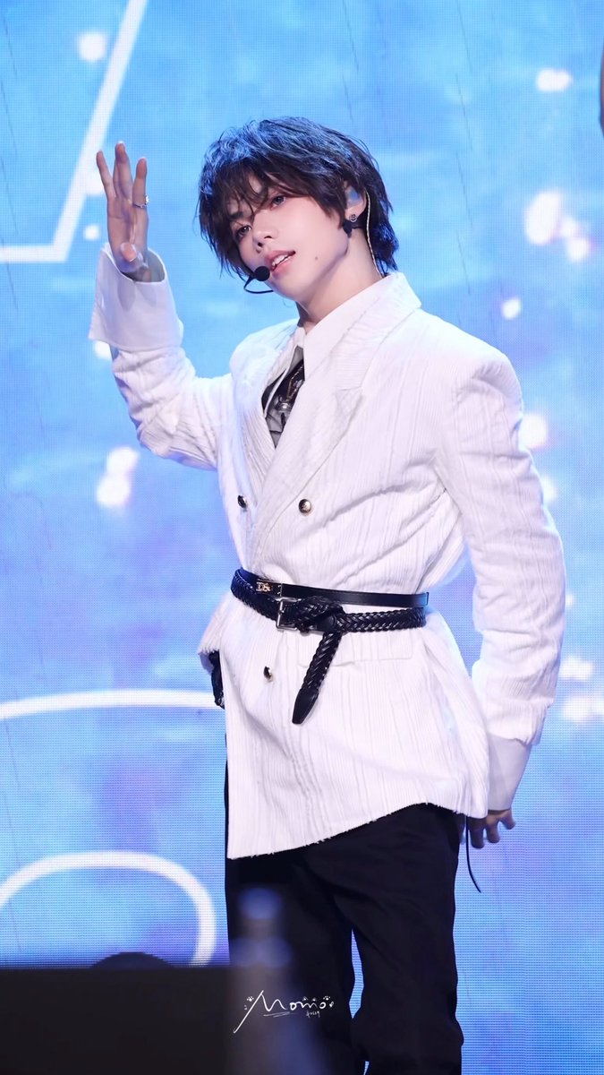 A young male performer with dark hair, wearing a white blazer over a shirt and tie with a braided black belt, poses on a stage illuminated by blue lighting with an in-ear microphone.