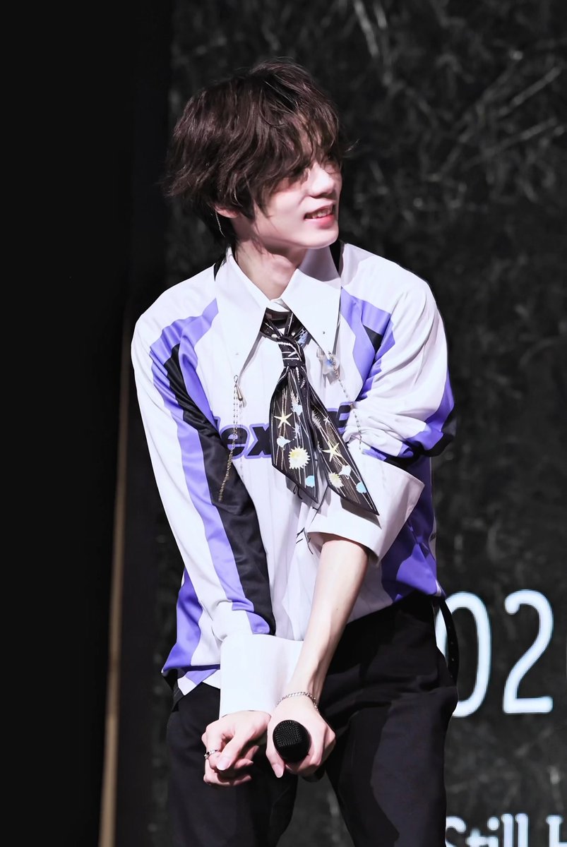A young man with wavy brown hair wearing a white and purple patterned shirt and black tie holds a microphone on stage, smiling to the side.