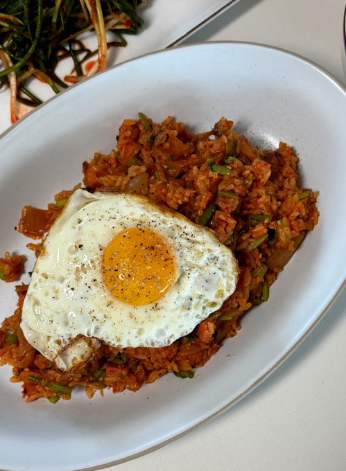 A plate of kimchi fried rice topped with a sunny-side-up fried egg seasoned with black pepper, served alongside a side of seasoned greens.
