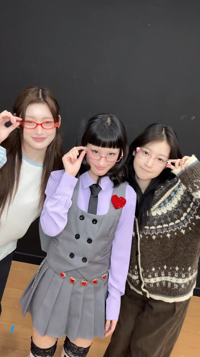 Three young women posing together in a room with dark walls, all adjusting small glasses, with the middle one wearing a purple shirt and grey vest outfit with a red heart pin.
