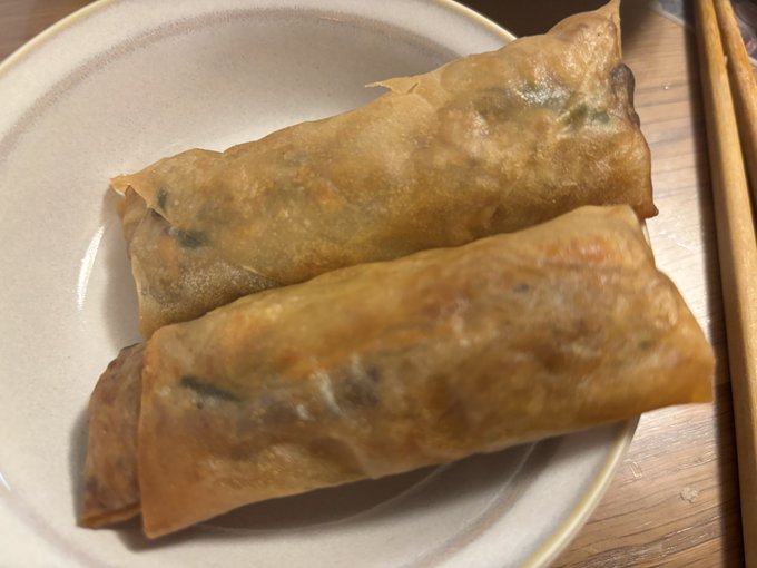 Two golden-brown fried spring rolls sit on a white plate next to wooden chopsticks on a wooden table.