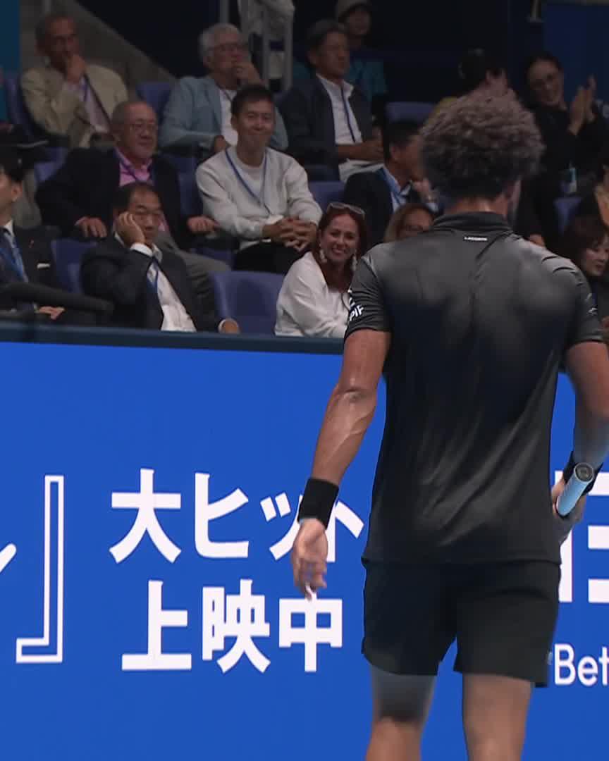 A tennis player with curly hair, seen from behind in a black athletic outfit, holds his racket while facing the crowd of spectators at a tournament. A blue barrier displays Japanese text "大ヒット上映中" and "Bet" branding, consistent with the Japan Open Tennis setting referenced in the post.