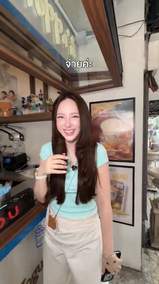 A candid-style photo of a smiling young woman with long dark hair wearing a light blue top and beige skirt, standing in what appears to be a shop, with the Thai text 'จ่ายค่ะ' floating above.