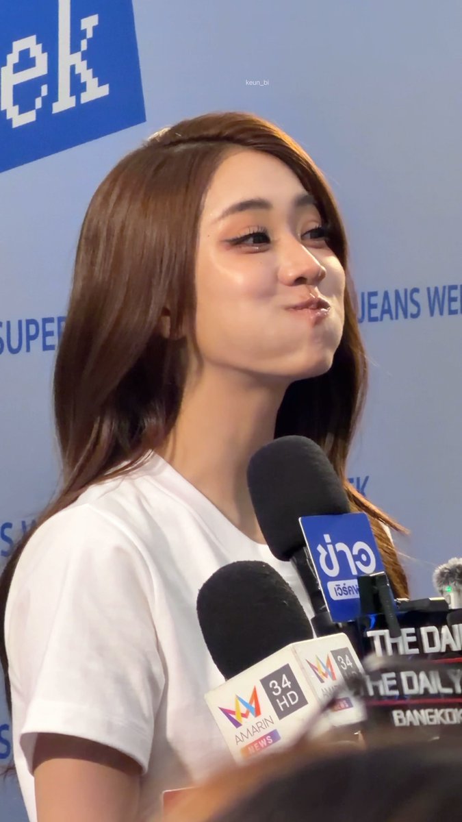 A young woman with long brown hair in a white t-shirt pouts playfully while speaking into multiple microphones from news outlets including Channel 8, Amarin 34 HD, and The Daily Bangkok.