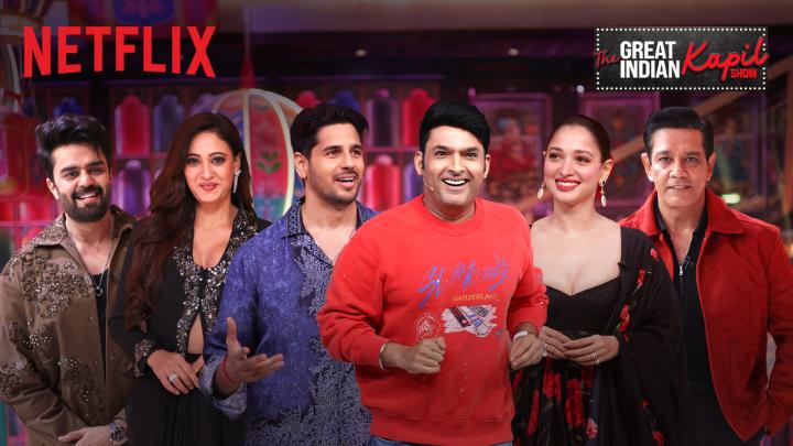 The image is a promotional poster for "The Great Indian Kapil Show" on Netflix, featuring six cast members including Kapil Sharma in a red sweatshirt at the center, alongside other smiling celebrities. The caption suggests a humorous moment on the show involving "Hansi" (laughter), tying the promotional image to the comedic and laughter-filled nature of the program.