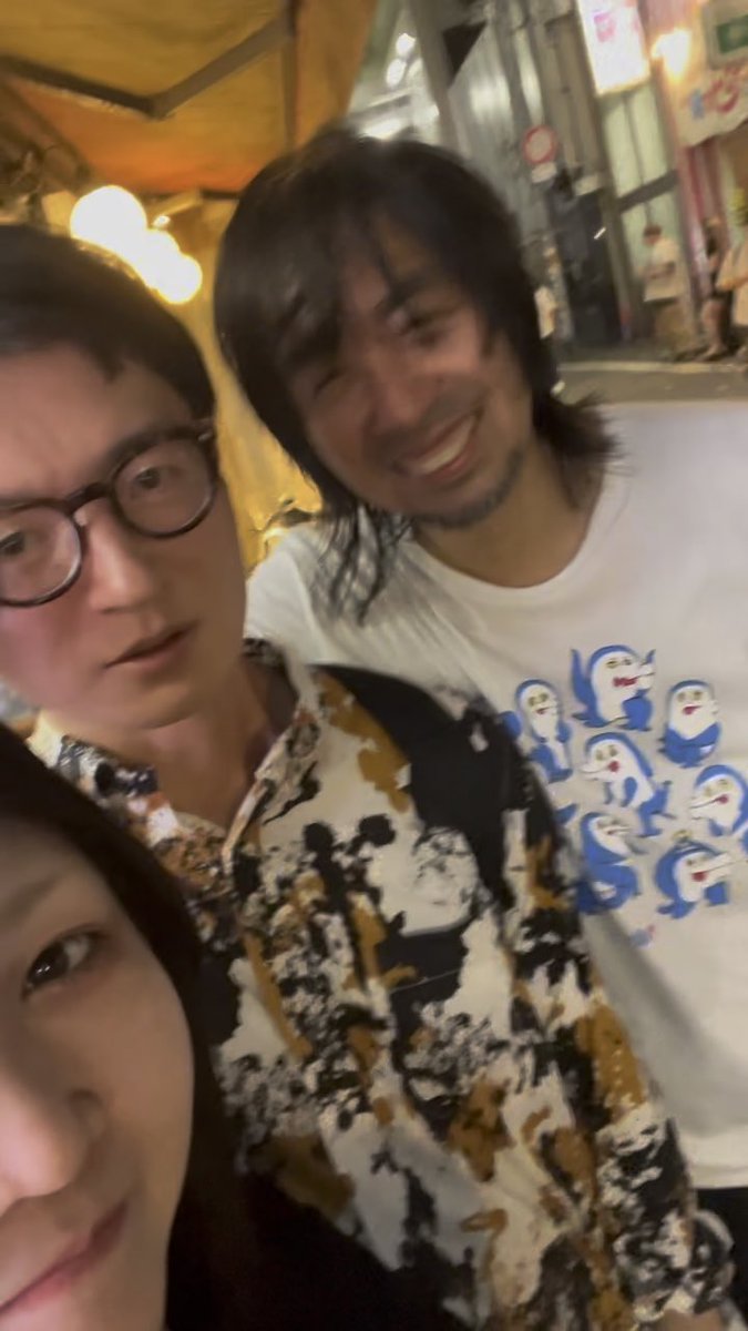 A nighttime selfie showing three people—a woman in the foreground, a man with glasses in a patterned shirt, and a bearded man in a white T-shirt featuring cartoon penguins—posing on what appears to be a city street.