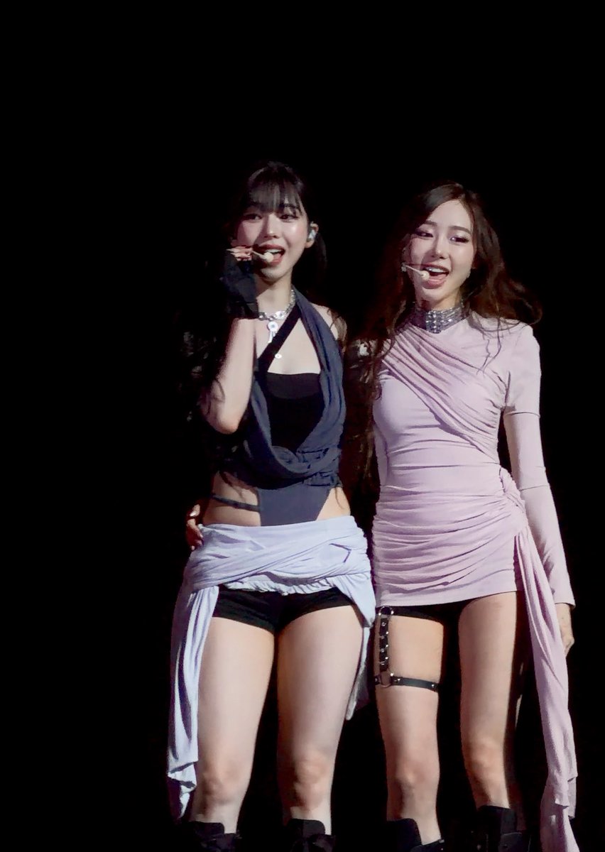 Two women perform on a dark stage, one in a black halter outfit with a white wrap, and the other in a long-sleeved pink dress with a studded choker.