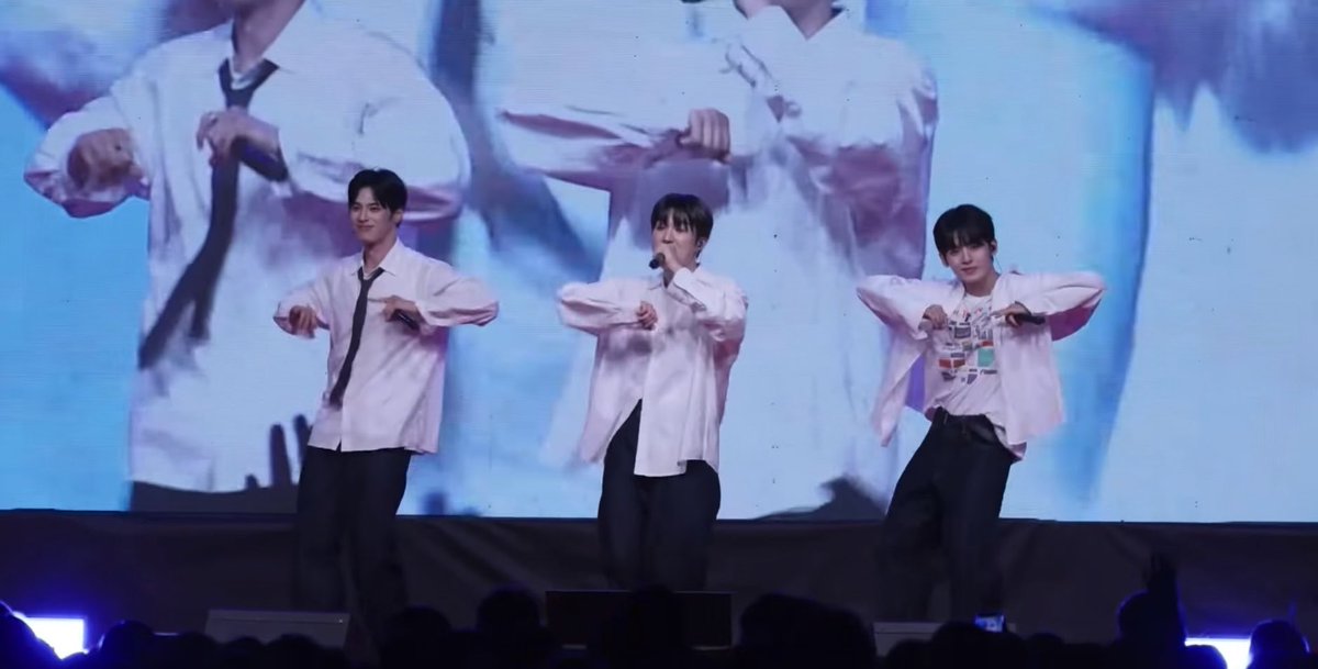 Three male performers in white shirts and black ties/pants performing choreography on a stage in front of a large screen, with audience silhouettes visible at the bottom.