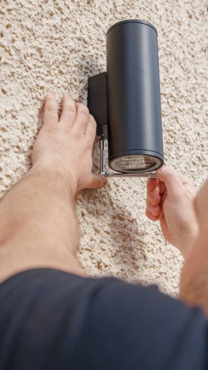 A close-up view showing hands using a tool to open a black cylindrical lockbox mounted on a textured wall, with a bare foot visible nearby.