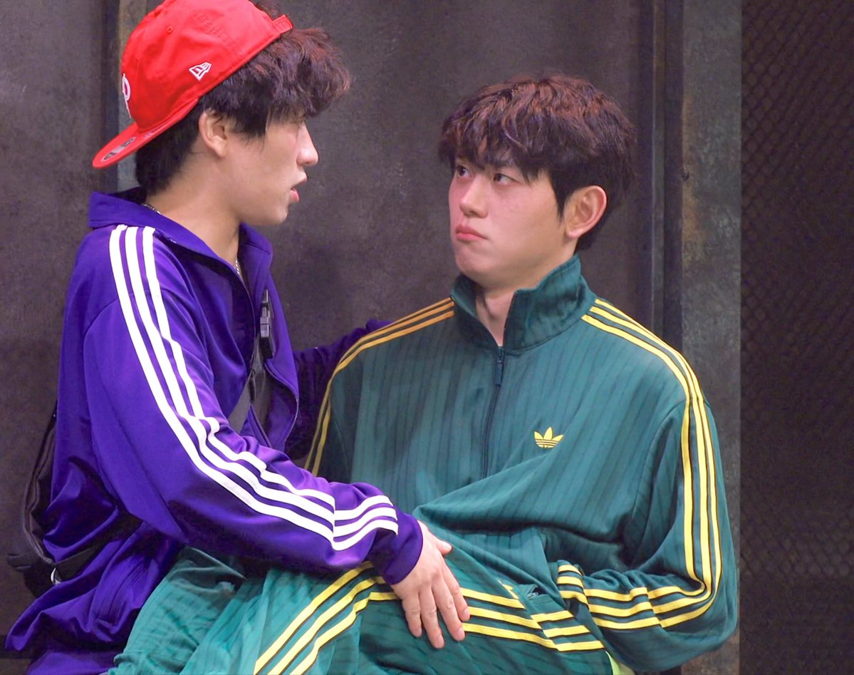 Two young men share a playful moment against a textured industrial backdrop—one in a purple Adidas track jacket and red New Era cap leaning in close to the other, who wears a green and yellow Adidas tracksuit and gazes back with a slightly annoyed expression. The scene captures the comedic dynamic referenced in the caption, where one is reassuring the other "Don't worry, we have Steve so we're good," tagging #안지환 (Ahn Jihwan) and #임태현 (Lim Taehyun), likely a humorous behind-the-scenes moment featuring these two Korean personalities.