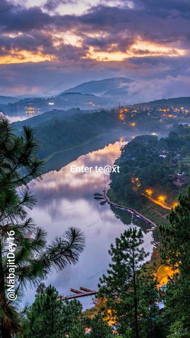 A scenic vertical wallpaper showing a sunset or sunrise over a misty river valley with scattered lights and boats, watermarked with '@NabaiTDeye' and 'Enter text'.