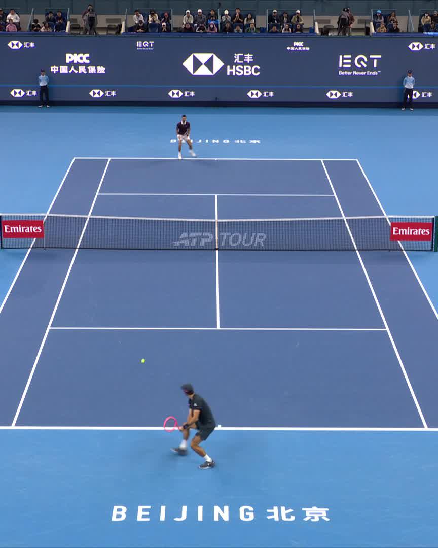Two tennis players compete on a blue hard court at the China Open in Beijing, with the ATP Tour net dividing them and sponsor logos from HSBC, PICC, EQT, and Emirates visible on the backdrop, while the player in the foreground prepares to return a ball during a long rally.