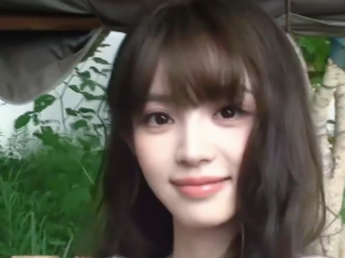 A close-up selfie of a young woman with long dark hair and bangs, smiling slightly with greenery visible in the background.