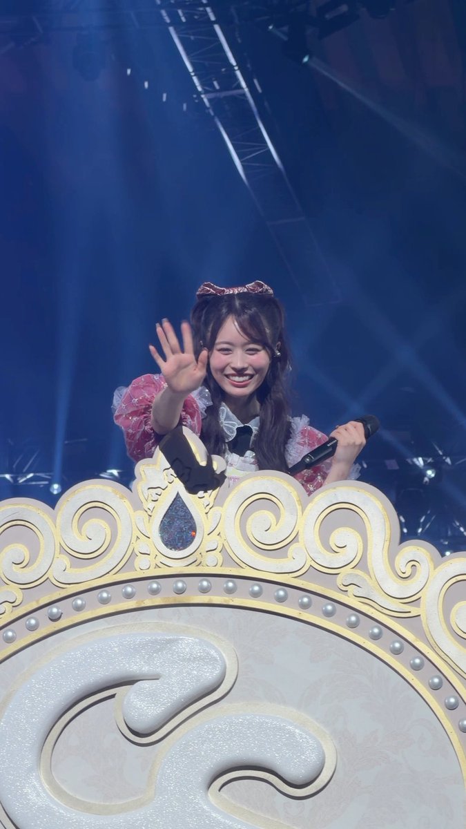 Image: A photo of a smiling young woman with long brown hair wearing a pink and white outfit, waving at the camera while holding a microphone, standing behind an ornate golden and white decorative stage element under blue stage lighting.