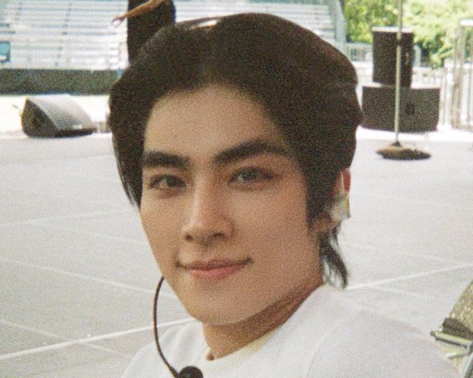 A close-up portrait of a young man with dark hair, wearing a white shirt and an in-ear monitor, likely a performer on stage.