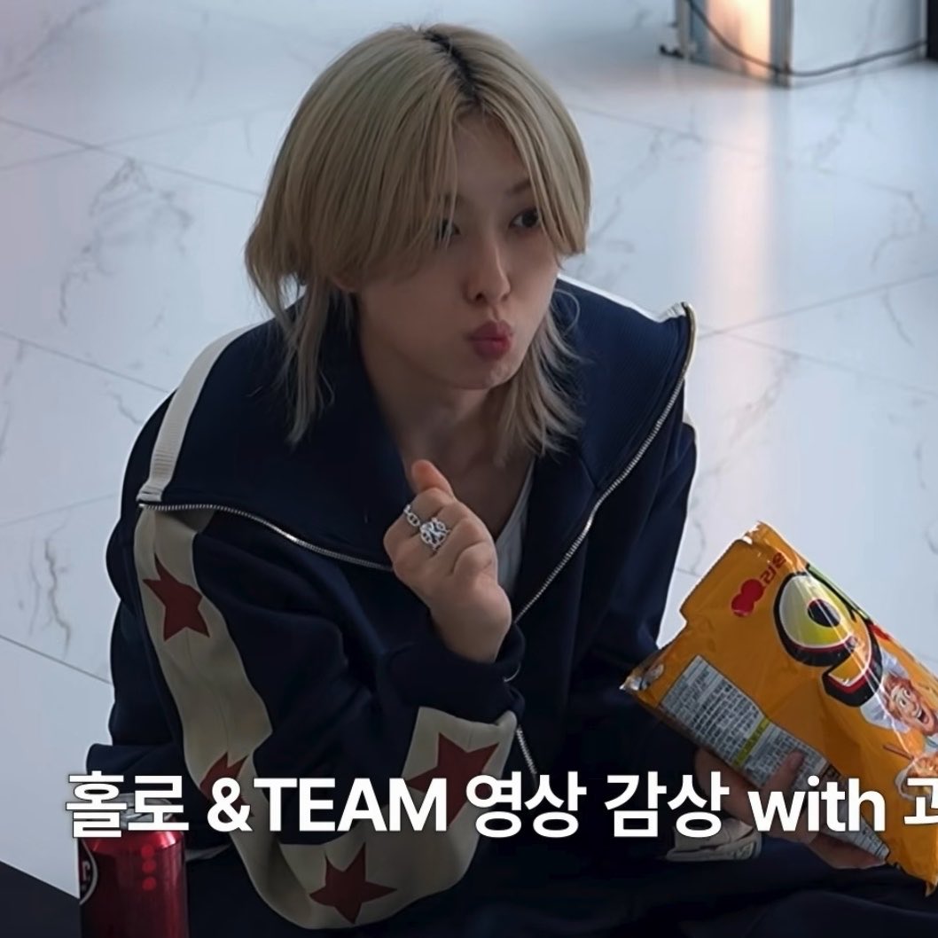 A person with blonde hair wearing a dark jacket with star patches sits on the floor holding a snack bag and making a finger heart gesture; Korean text reads "홀로 &TEAM 영상 감상 with" (Watching Holo &TEAM video with).